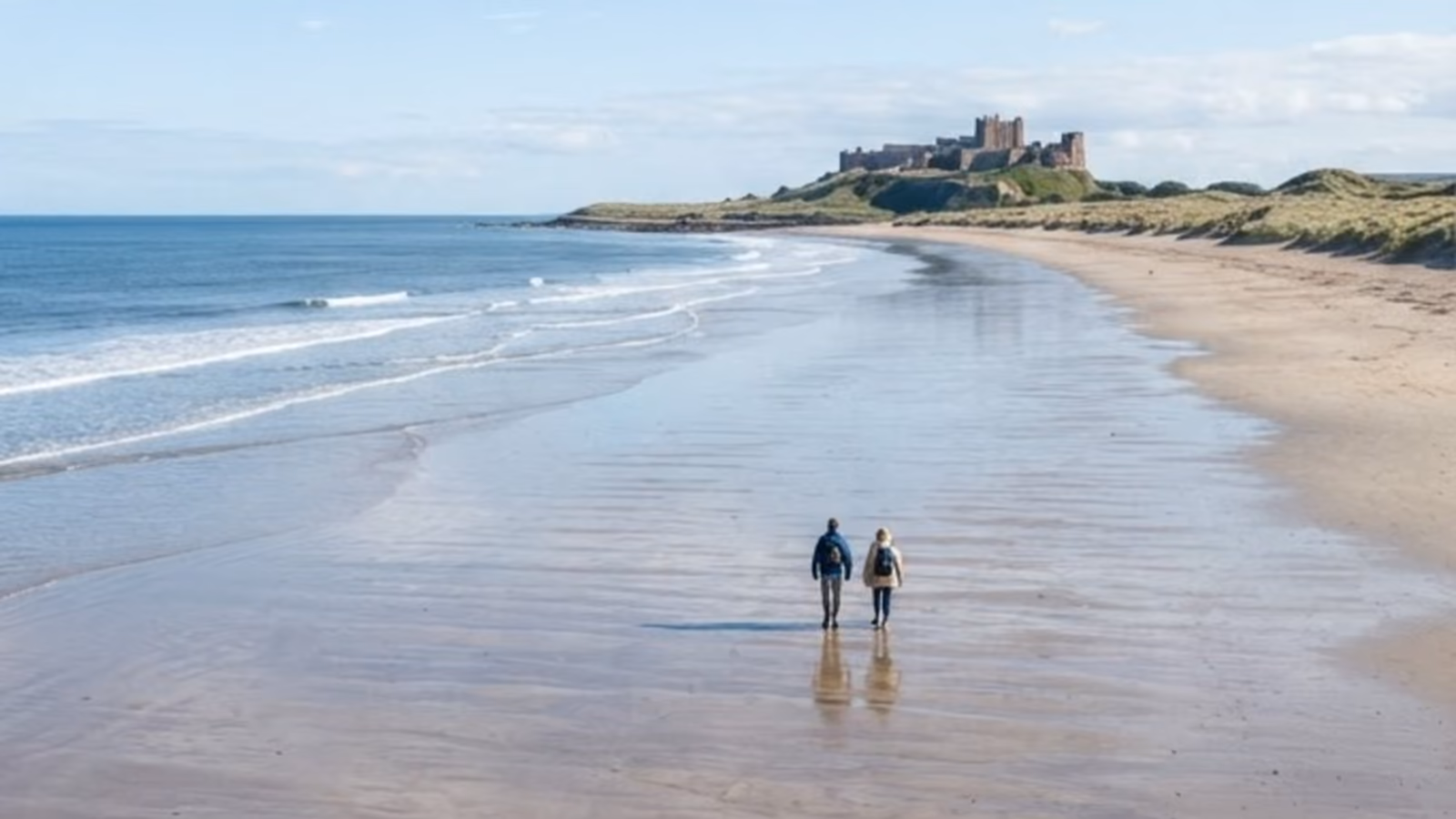 Two people walking the wet sand of a wide empty beach, a castle standing on the dunes ahead of them