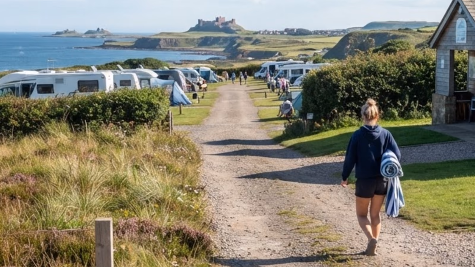 A woman carrying a rolled towel walks the gravel track past the shower block, motorhomes and a castle headland beyond