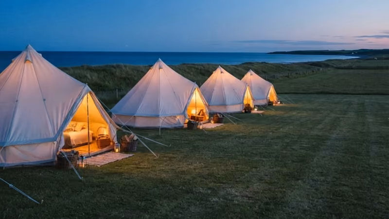 Bell tents glowing along the cliff edge at dusk, the sea beyond