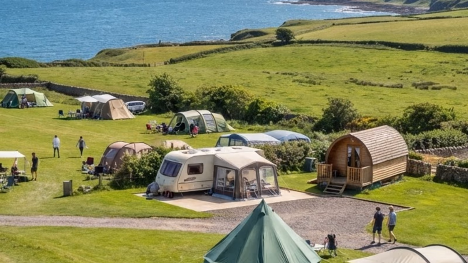 Tents, a caravan under an awning and a timber glamping pod spread across a green field above the sea